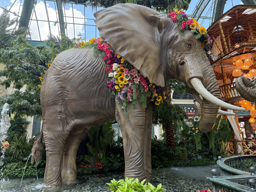 Large elephant sculpture draped with colorful flowers, standing in a pool beneath the glass roof of the Bellagio Conservatory.
