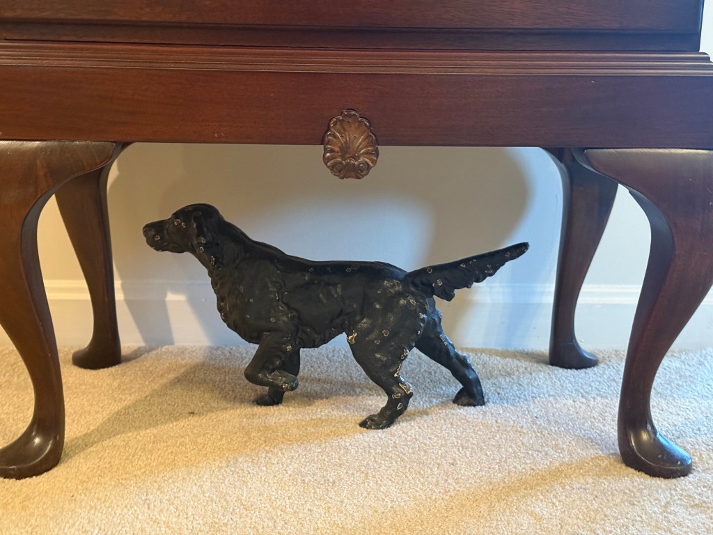 Black iron pointer dog with a worn finish standing beneath my mother’s writing desk, framed by its curved legs.