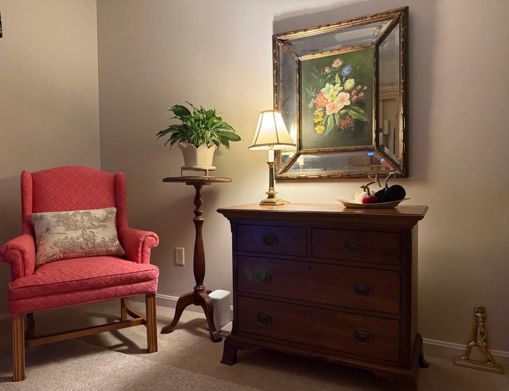 Floral painting above a chest of drawers, illuminated by a brass table lamp, with a pink wingback chair and potted plant beside it.
