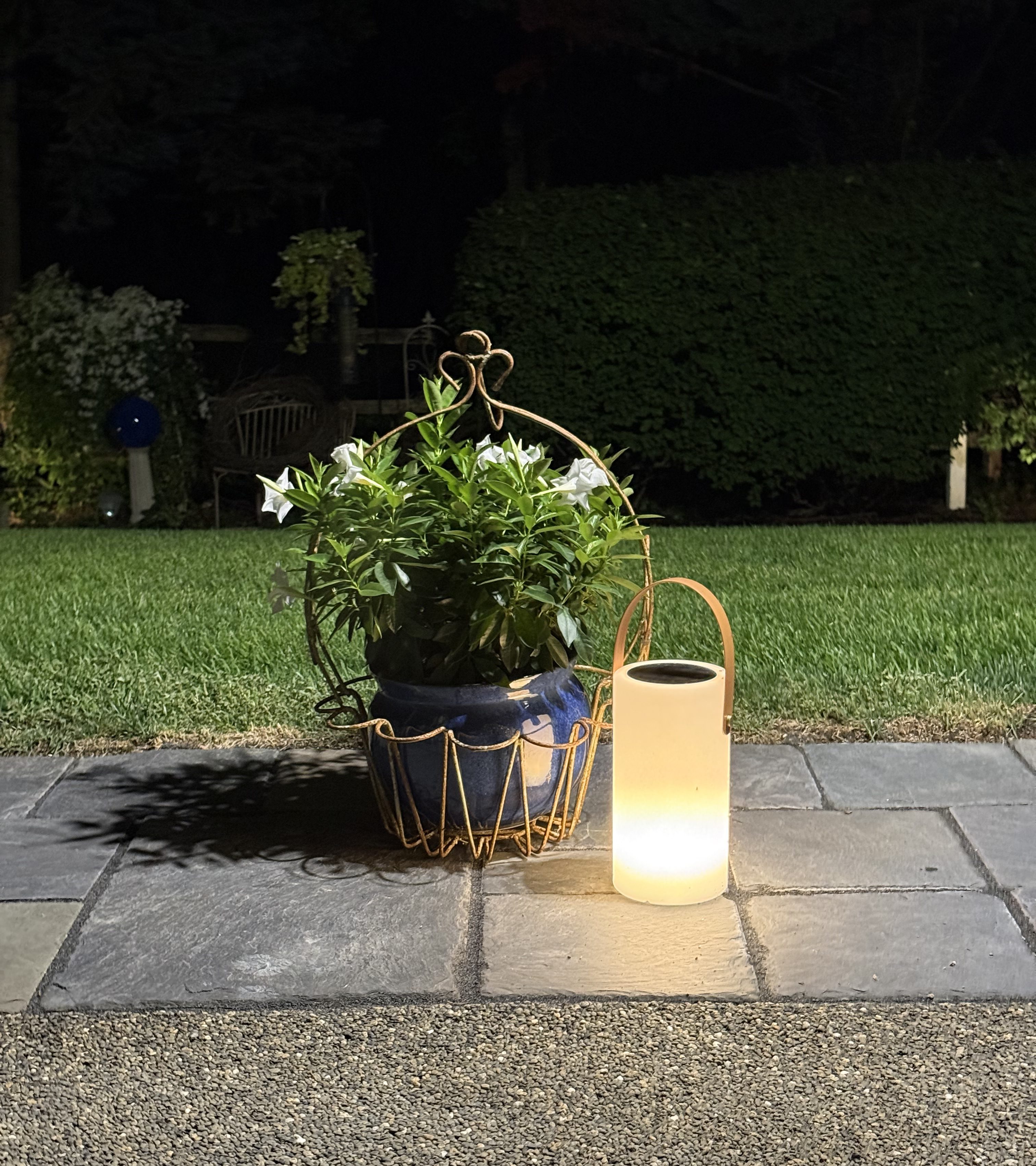 Solar lantern on its white setting beside a blue pot of white flowers on the patio at night.