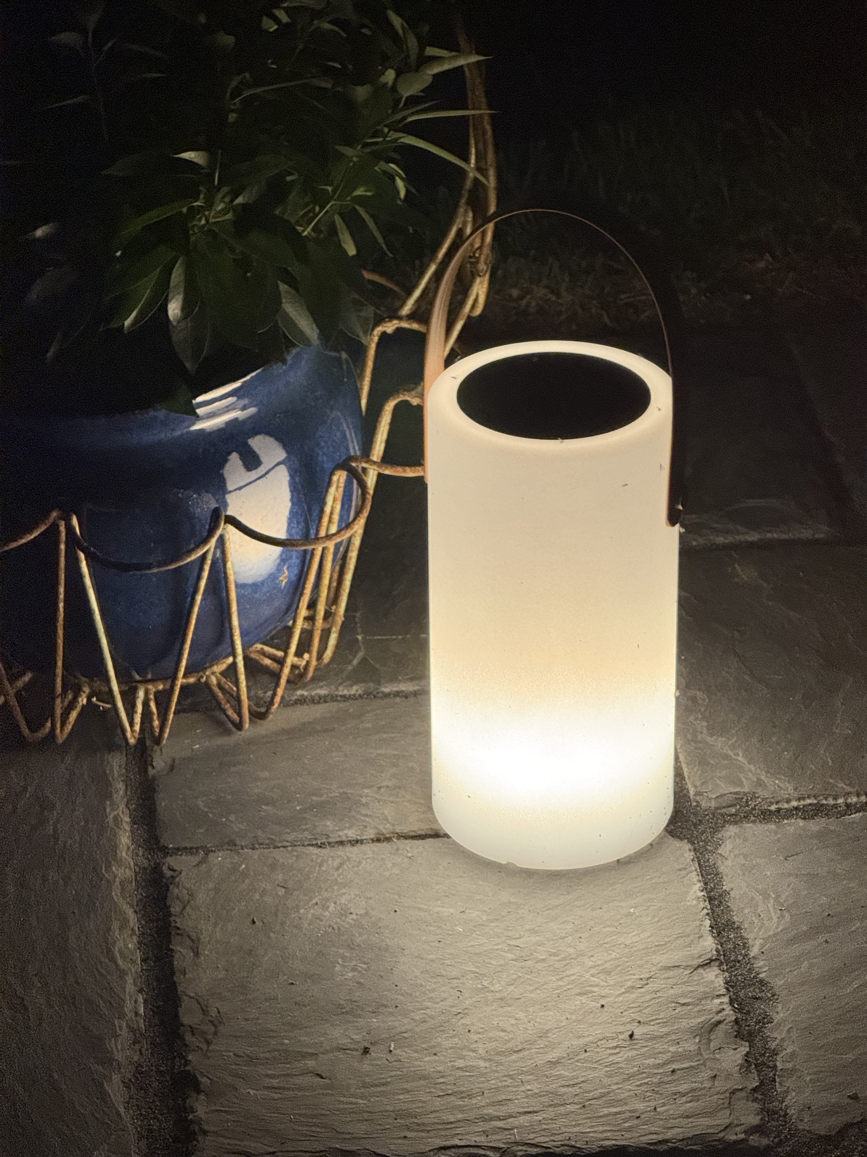 Close-up of a glowing solar lantern on its white setting beside a blue planter on the patio at night.
