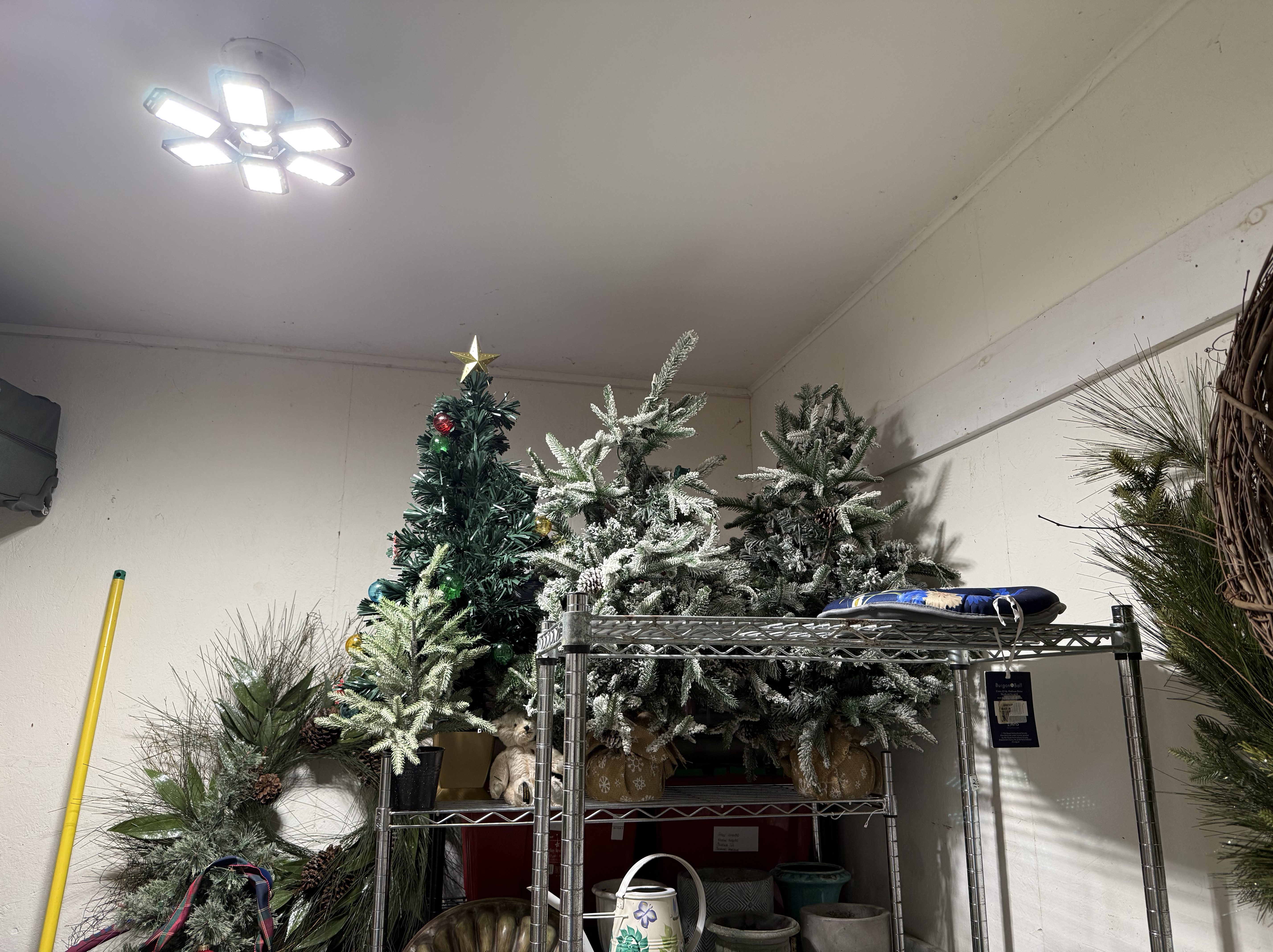 LED ceiling light illuminating storage shelves with artificial Christmas trees, planters, and garden decorations.
